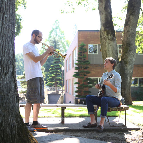 Two students play a banjo and trumpet outside on the quad