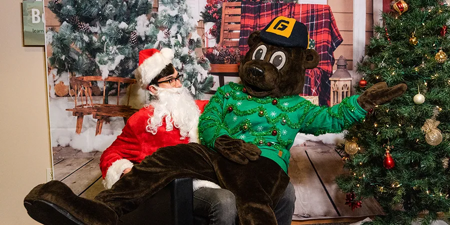 Mascot Pennington sitting on Santa's lap wearing a Christmas sweater