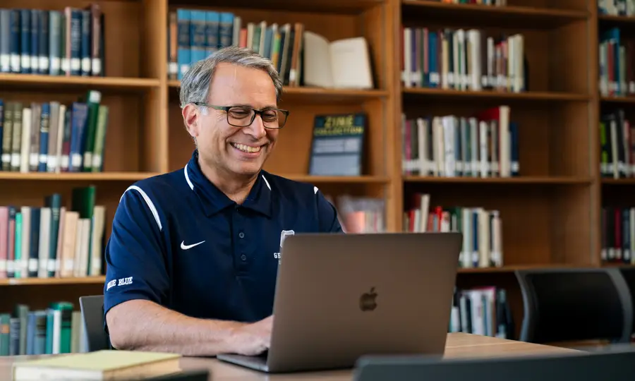 Rick Muthiah wearing a George Fox polo smiling at a computer sitting in front of a wall of books