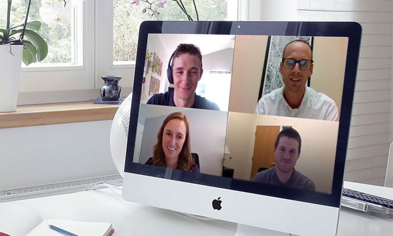 Photo of a computer screen on a desk. The screen shows a video call with four people.