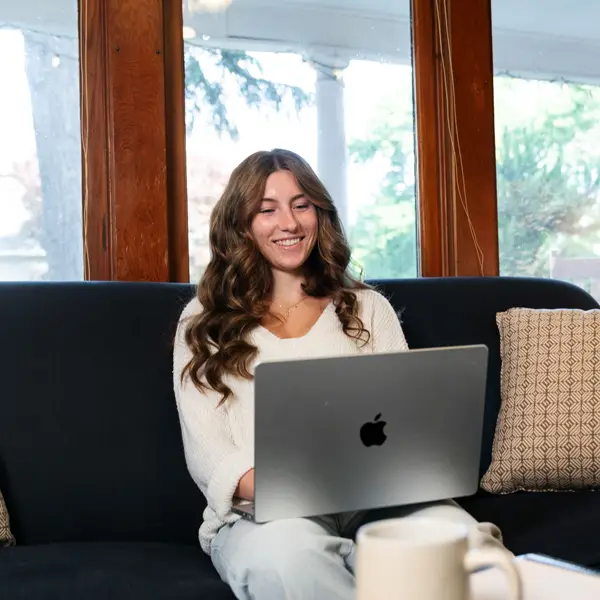 A girl sitting on her couch in front of a window smiling at her laptop