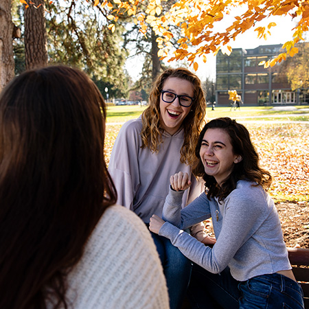 Three students sitting on a bench on campus laughing together