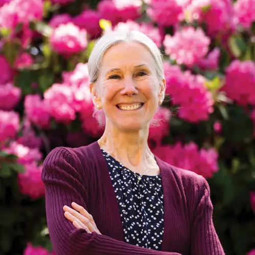 MaryKate Morse smiling with her arms crossed in front of a full bush of rhododendrons