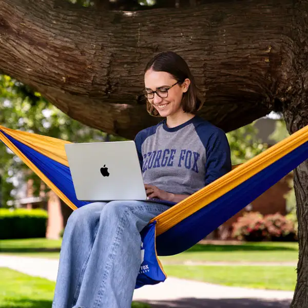 A student wearing a George Fox shirt and sitting in a hammock working on her computer
