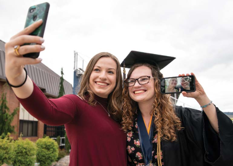 Two women, one is taking a selfie of both of the scene while the other, in graduation robes, is holding up a phone video calling two other women
