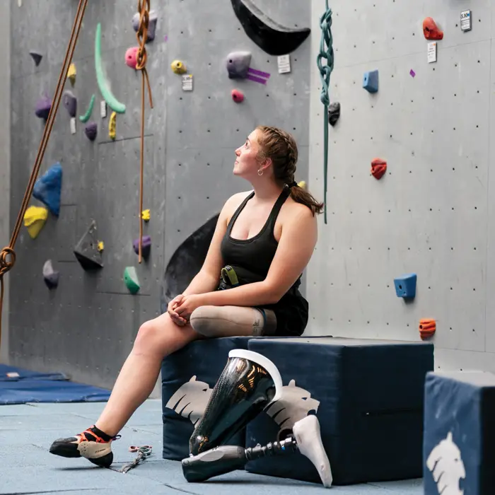 Isabelle sitting on a cube, smiling up at the wall, ready to climb with her prosthetic leg set beside her