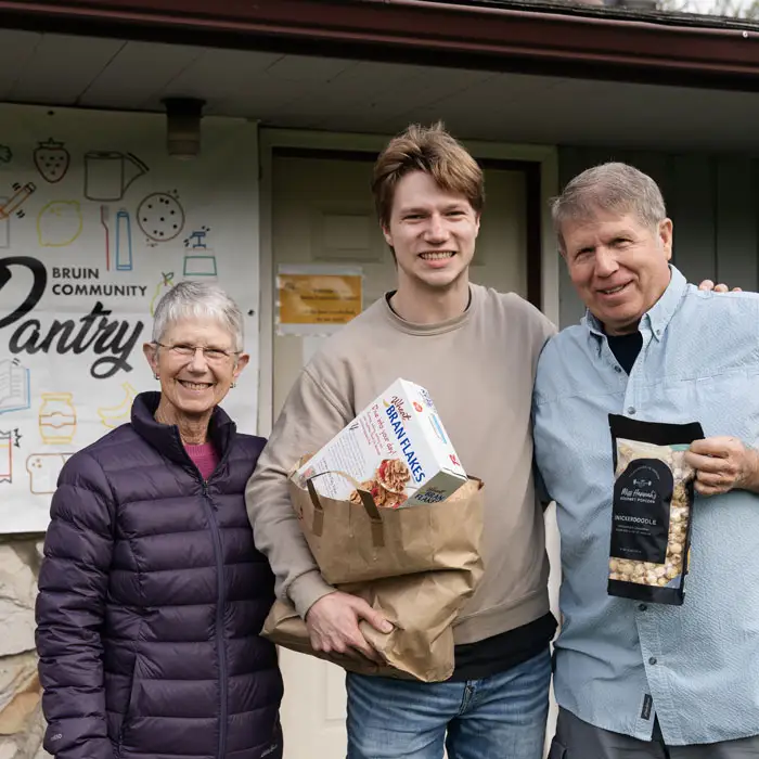 A student standing in front of the Bruin Community Pantry with two others and holding a bag of food