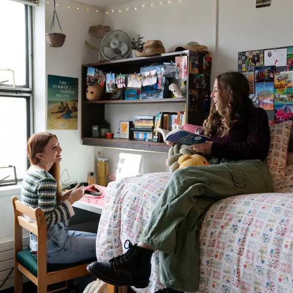 Student doing her makeup in her dorm vanity while her roommate reads a book on the bed next to her