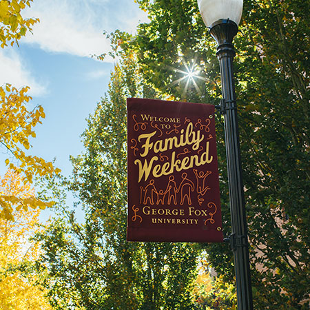 Lamppost banner that reads 'Welcome to Family Weekend, George Fox University' with trees and sun behind