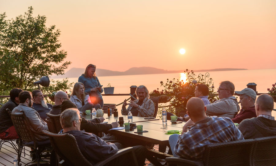 people sit outside at a table while the sun sets over a lake